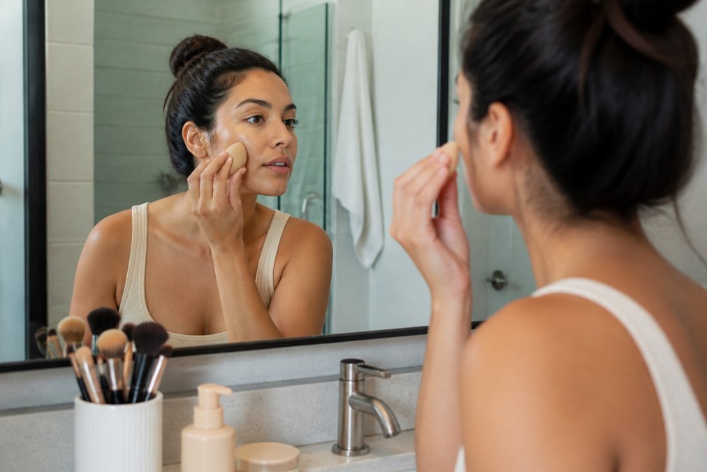 Woman with medium skin tone applying natural makeup in a bright bathroom mirror, showing foundation application with a beauty sponge and brushes nearby on the vanity