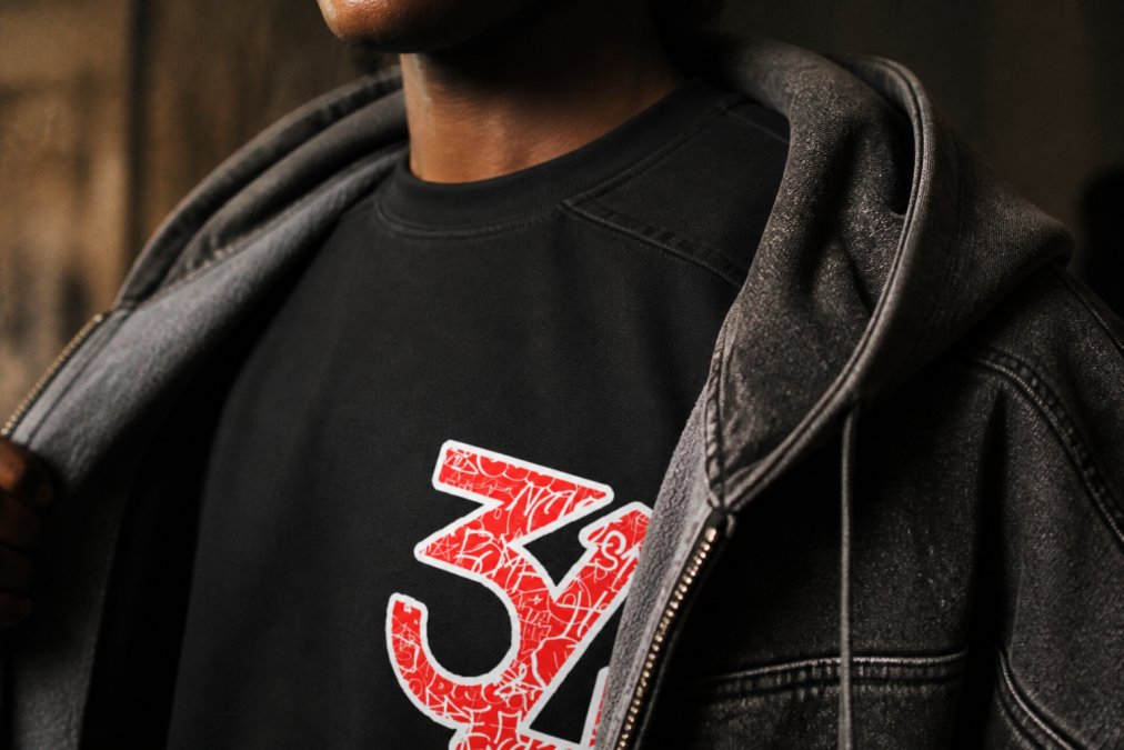 Close-up detail shot showing layered urban fashion with visible raw seam paneling on premium cotton tee, distressed snow-washed fabric texture on overlying hoodie, and visible stitching detail demonstrating fabric quality and craftsmanship under warm studio lighting