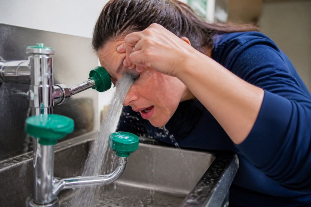 Person rinsing their eye at a sink or eyewash station with clean running water, demonstrating proper first aid technique with their head tilted and eye held open under the stream