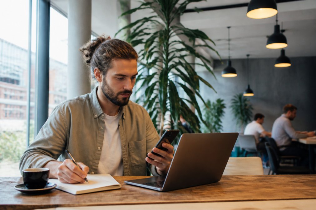 Digitaler Nomade arbeitet konzentriert an Laptop und Smartphone in hellem Co-Working-Space oder modernem Café, mit Notizbuch und Kaffee, symbolisiert ortsunabhängige Gründung einer US LLC
