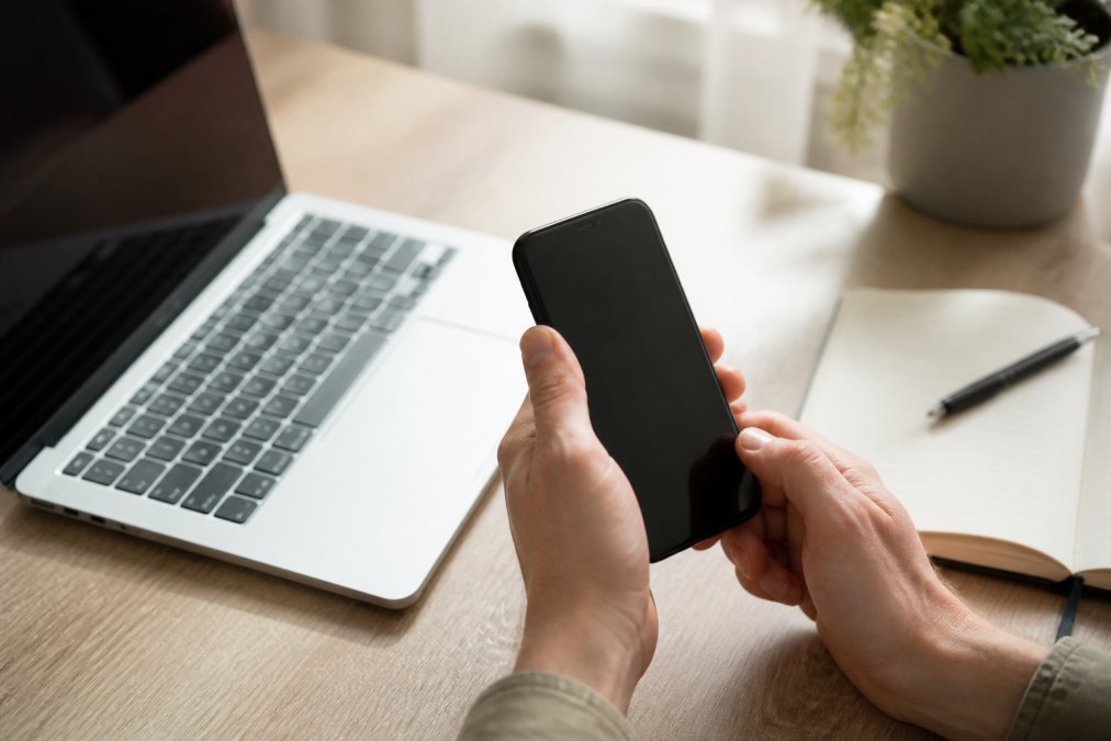 Nahaufnahme von Händen, die ein Smartphone halten und eine Banking-App öffnen, Laptop und aufgeschlagener Notizblock im Hintergrund auf einem hellen Holzschreibtisch, weiches Tageslicht