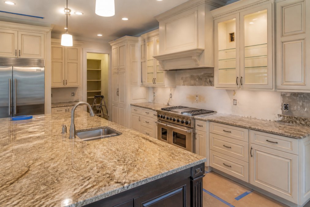 Luxury kitchen renovation in progress with custom cabinetry, granite countertops, and stainless steel appliances under bright work lights, showing detailed craftsmanship and attention to detail