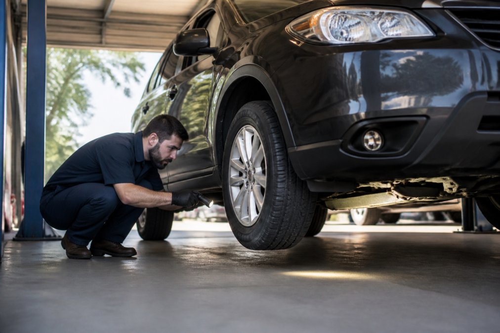 Professional mechanic crouching beside a used SUV, using a flashlight to examine the undercarriage and tires during a detailed pre-purchase inspection in daylight