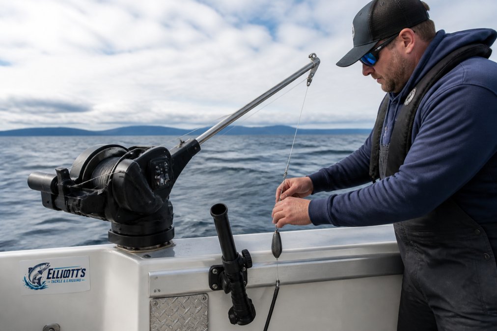 Professional angler adjusting an electric downrigger mounted on a modern saltwater fishing boat with stainless steel boom extended, showing the rod holder and release clip system during a halibut trolling session on the open water