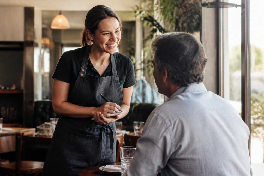 Friendly server using guest data to greet a regular customer by name in a cozy, sunlit restaurant dining room.