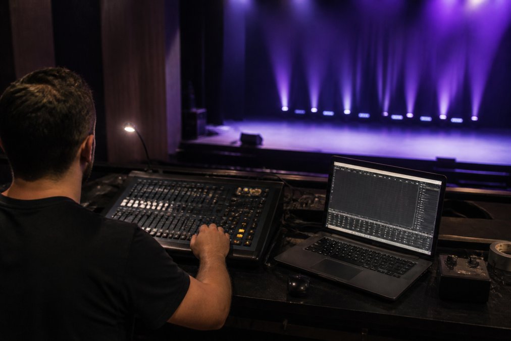 A lighting technician at a small theater console, laptop open beside the desk showing a patch window, smart LED fixtures glowing in the background on stage