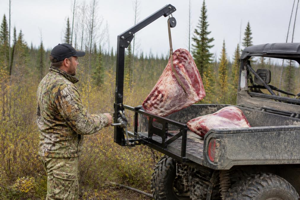 A hunter operating a UTV-mounted game hoist to lift a moose quarter onto a cargo rack in a rugged bush setting, showing the mechanical advantage and solo operation