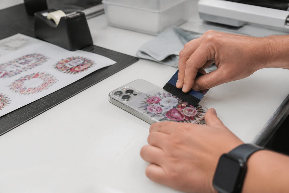 Close-up of hands applying a UV DTF transfer sticker to a smartphone case with a squeegee on a clean workbench
