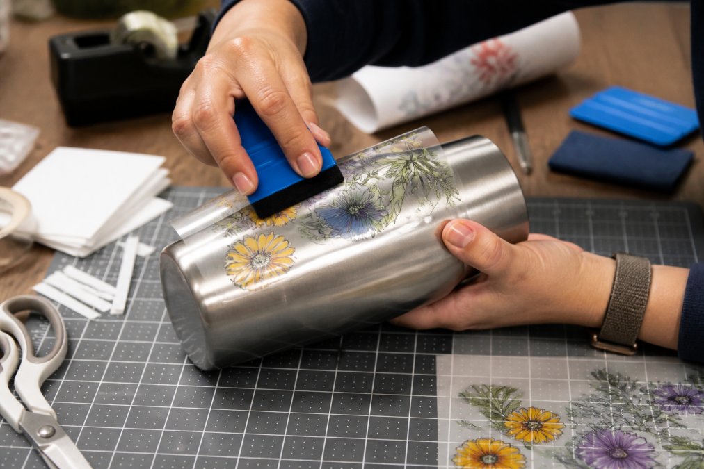 Close-up of hands applying a UV DTF transfer to a stainless steel tumbler using a squeegee, with a small craft workspace visible in the background