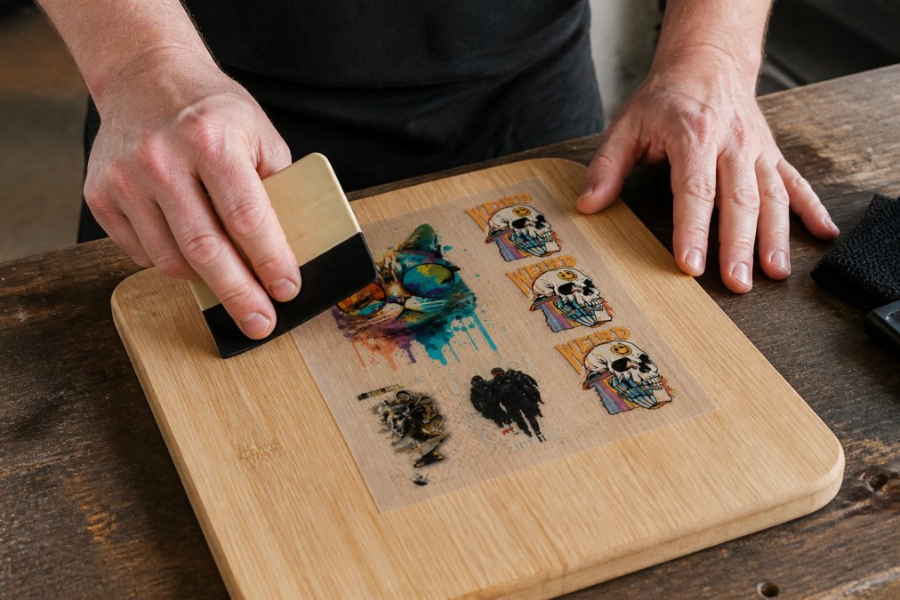 Close-up of hands carefully positioning a UV DTF transfer onto a wooden surface, using a squeegee tool to apply firm, even pressure from centre outward, with natural workshop lighting
