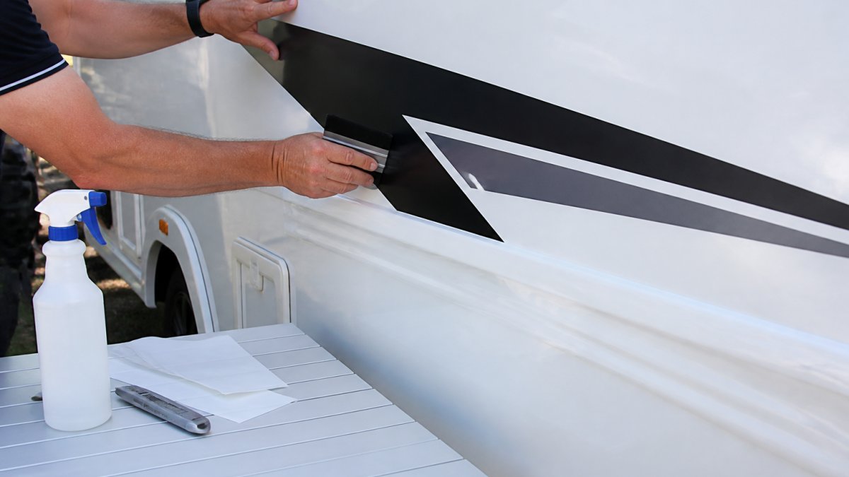 Close-up of a person's hands using a squeegee to smooth a large vinyl decal onto the flat side panel of a white caravan, with a spray bottle and trimming tool resting nearby on a folding table outdoors