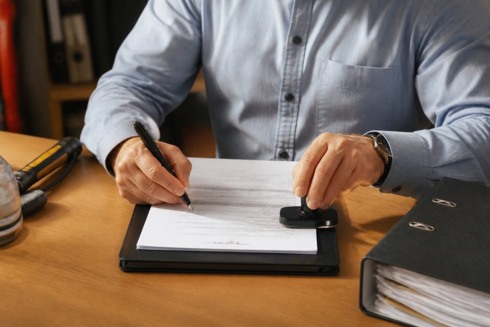 Nahaufnahme eines Arbeitssicherheitsexperten im Businesshemd, der an einem aufgeräumten Schreibtisch ein ausgefülltes Prüfprotokoll mit Stempel und Unterschrift abzeichnet, warmes Bürolicht