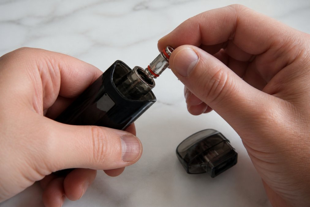Hands carefully inserting a replacement SMOK RPM coil into a Nord pod, showing the screw-in mechanism and proper handling technique to avoid damaging the coil pins, with a Uwell replacement pod visible nearby on a clean surface