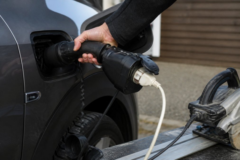 Närbild av en hand som ansluter en typ 2 V2L-adapter till en elbils laddningsport, med ett elverktyg inkopplat i Schuko-uttaget på adaptern.
