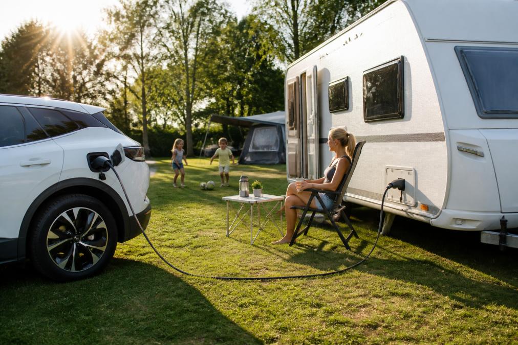 Familie auf dem Campingplatz mit Elektroauto und Wohnwagen, der über ein V2L-Kabel angeschlossen ist, Sonnenschein, spielende Kinder im Hintergrund
