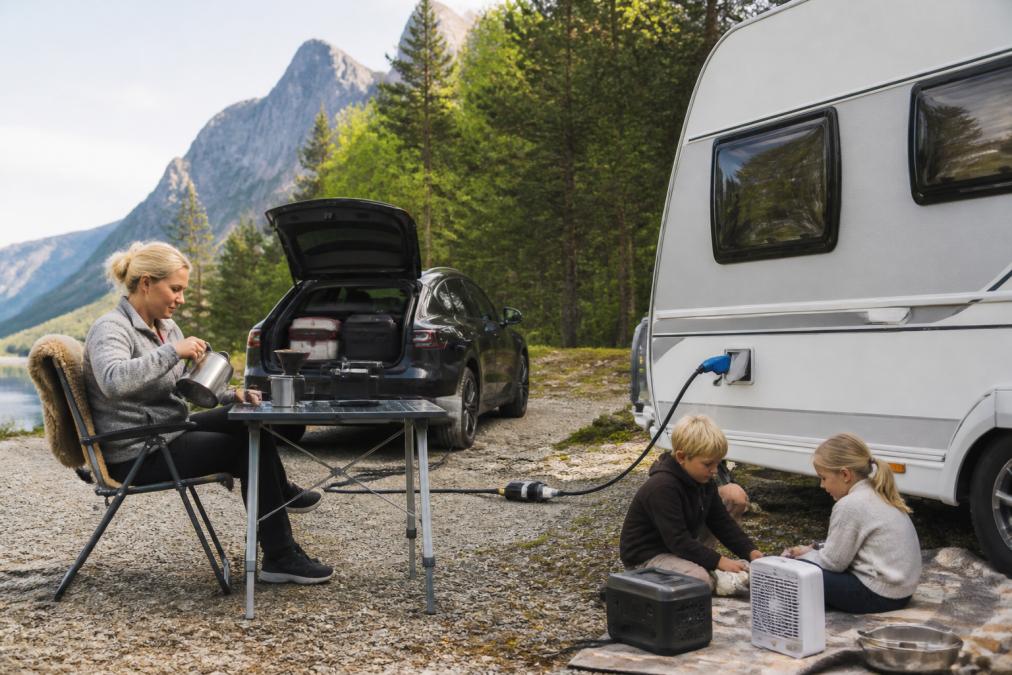 Familj med husvagn i naturlig omgivning, med elbil i bakgrunden; campare använder 230 V ström för utomhusapparater, vattenkokare och campingutrustning ansluten via typ 2-adapter