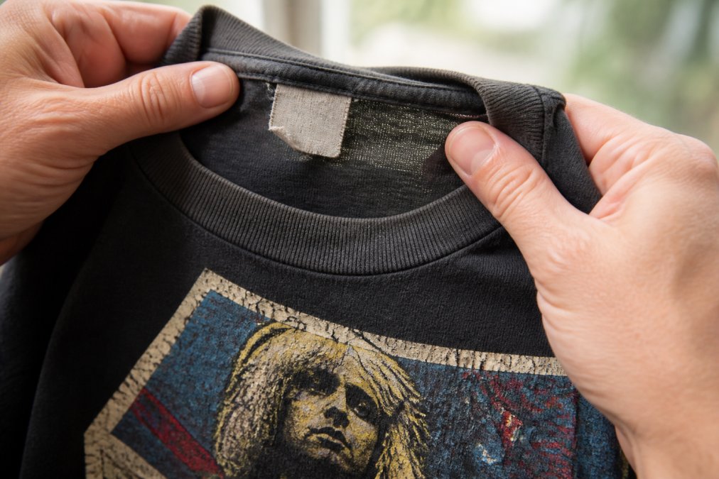 Close-up of hands holding a faded vintage concert t-shirt against natural light, showing the worn collar tag and cracked screen print on the front graphic