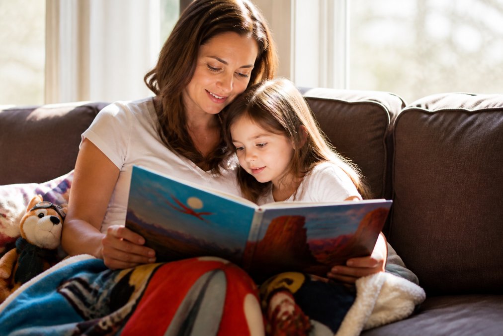 Parent and child sitting together on a comfortable couch reading an adventure-themed picture book, child leaning in with engaged expression, warm natural light from nearby window illuminating the pages