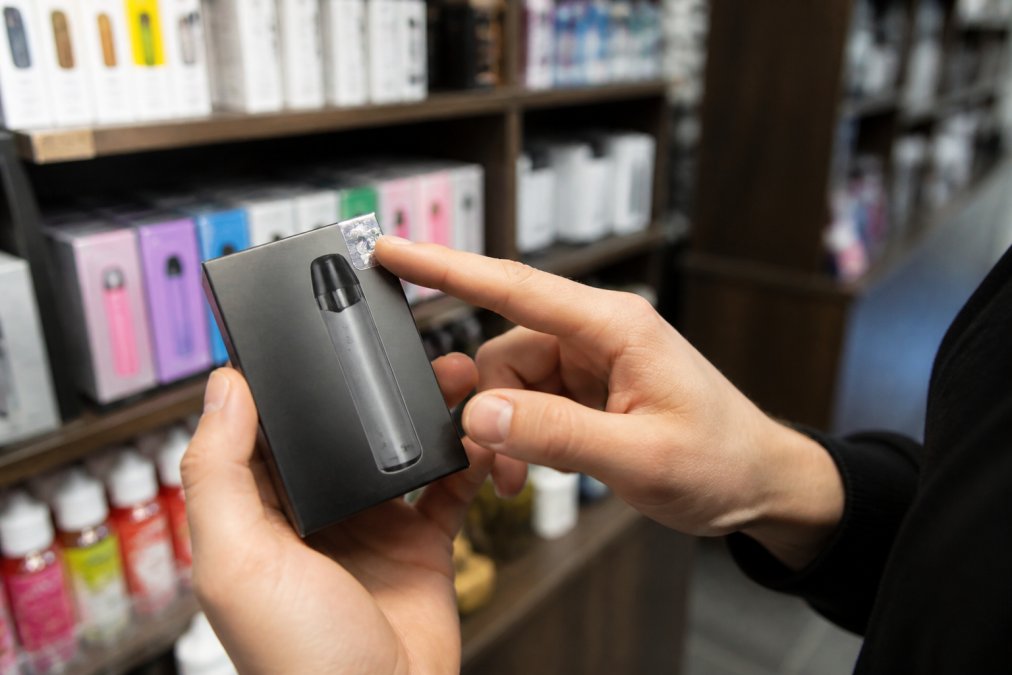 Close-up of a staff member's hands holding a branded vape device box, pointing to an authenticity hologram sticker under bright retail lighting in a well-organized shop