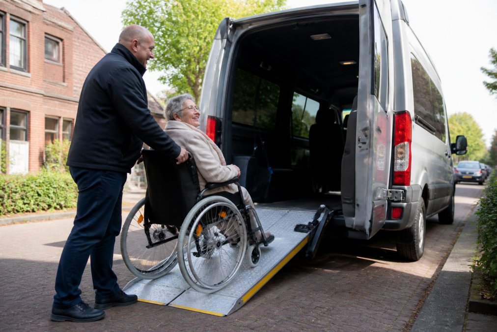 Gecertificeerde chauffeur helpt voorzichtig een oudere rolstoelgebruiker via een uitgeschoven oprijplank een rolstoeltoegankelijke taxibus in, beide personen glimlachen in een rustige woonstraat op een zonnige ochtend