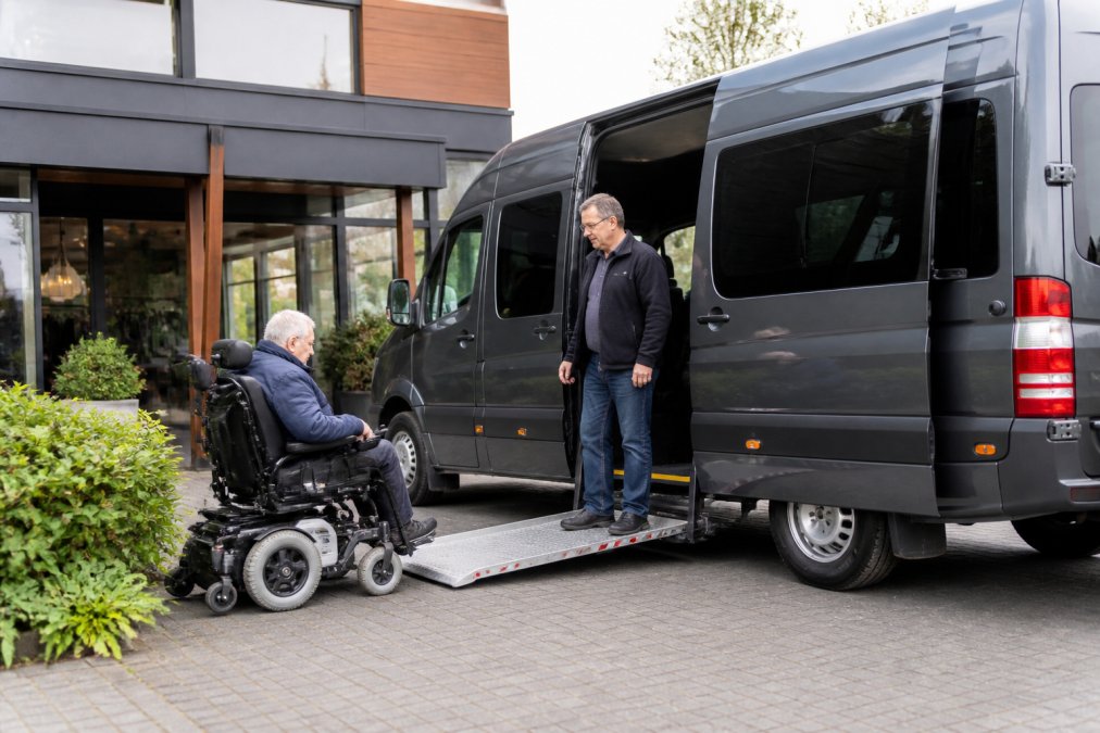 Rolstoeltoegankelijke taxibus geparkeerd voor de ingang van een dagbestedingscentrum, chauffeur staat bij de open schuifdeur met uitgeschoven oprijplank terwijl een passagier in een elektrische rolstoel de oprijplank nadert