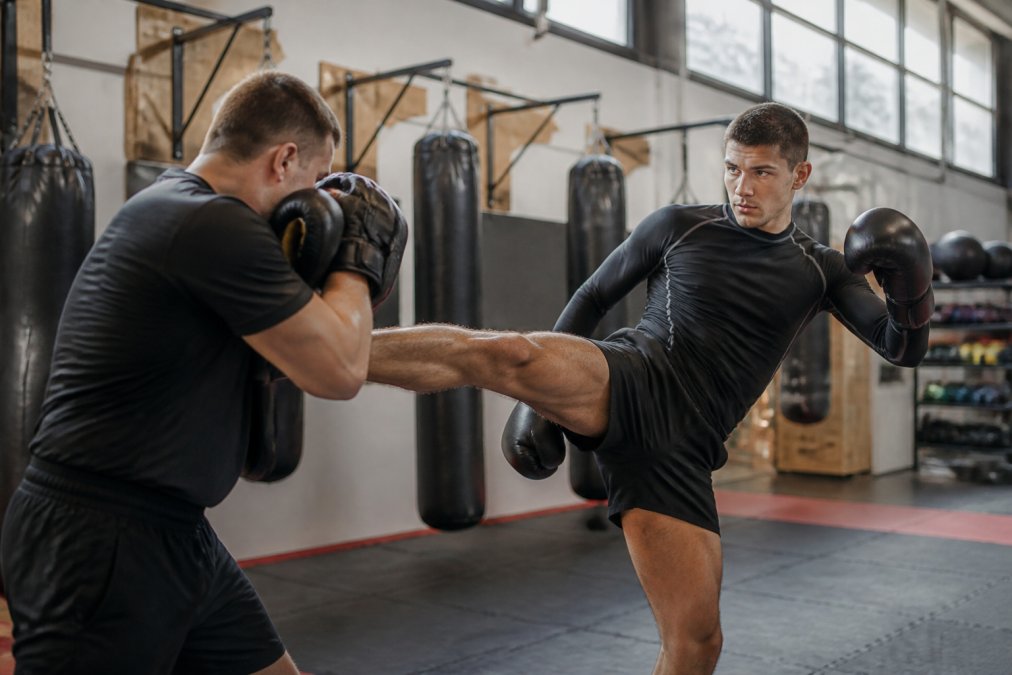Een vechtsporter in een rashguard en kickboksbroekje tijdens een training in een gym, close-up van de pasvorm en bewegingsvrijheid van de kleding, natuurlijk licht uit hoge ramen