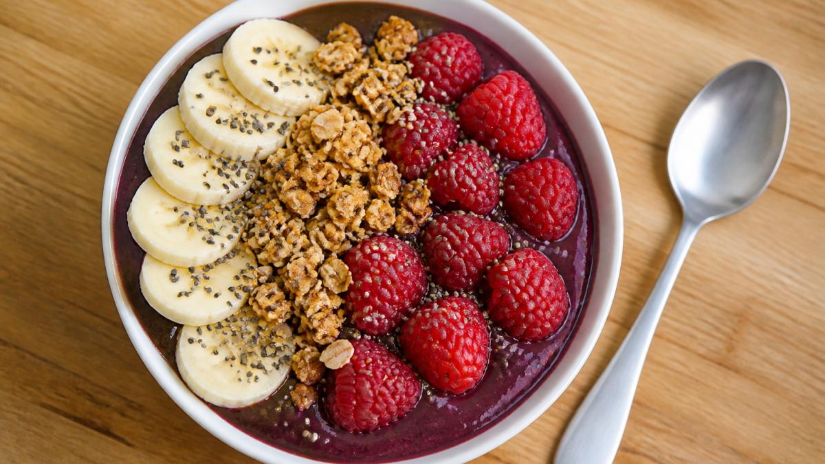 Overhead shot of a thick purple açaí bowl in a white ceramic bowl topped with sliced banana, raspberries, granola, and chia seeds, sitting on a light wood counter with a spoon resting beside it