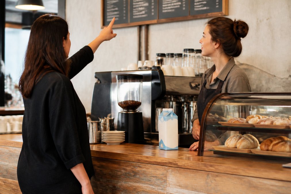 A customer at a coffee shop counter talking with a barista, pointing at a menu board, with a dairy-free milk carton visible on the counter behind the espresso machine