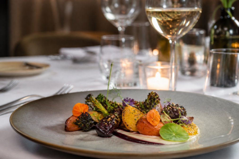 Elegantly plated plant-based tasting menu course featuring roasted seasonal vegetables, microgreens, edible flowers, and artistic sauce work on fine dining tableware, with wine glasses catching soft candlelight in background