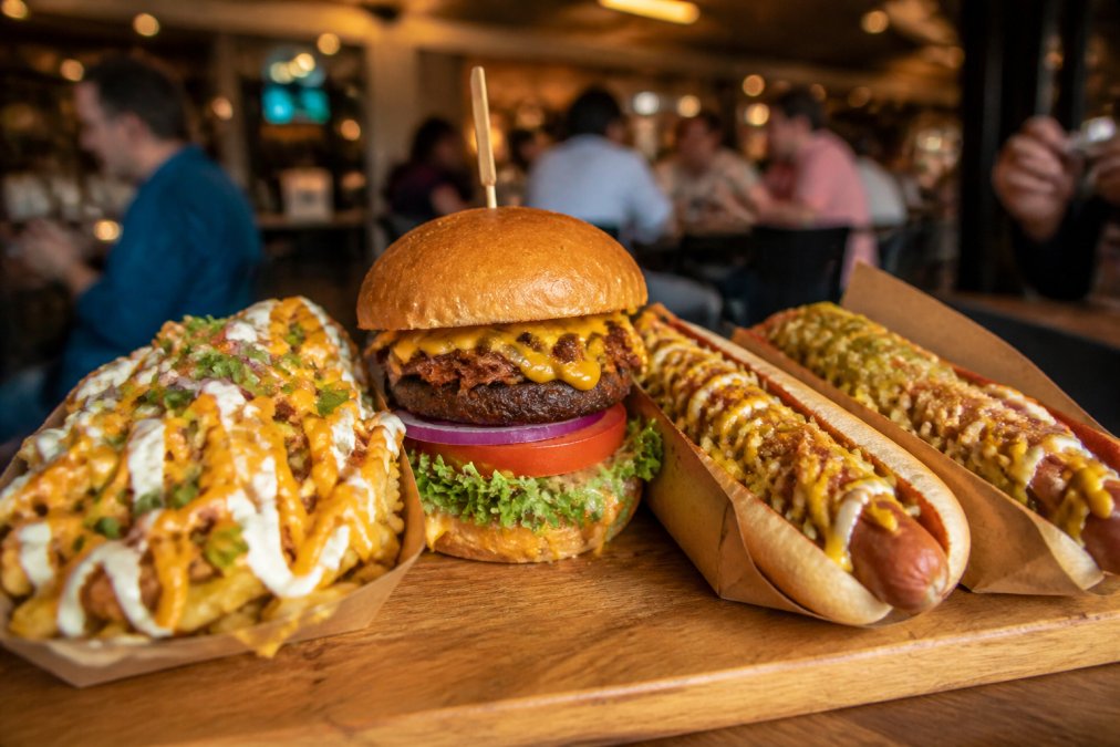 Close-up of indulgent vegan street food dishes, loaded plant-based burgers stacked with toppings, loaded fries piled high with vegan cheese and sauces, and vegan hot dogs, arranged on a wooden board in a casual market setting with warm overhead lighting and blurred diners in background