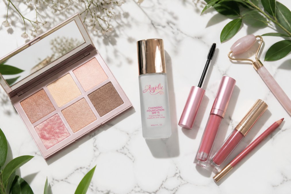 Flat lay of vegan makeup products including mascara, lip kits, and a highlighter palette arranged on a clean white marble surface with fresh botanical leaves and a small jade roller as accents, photographed in soft natural daylight