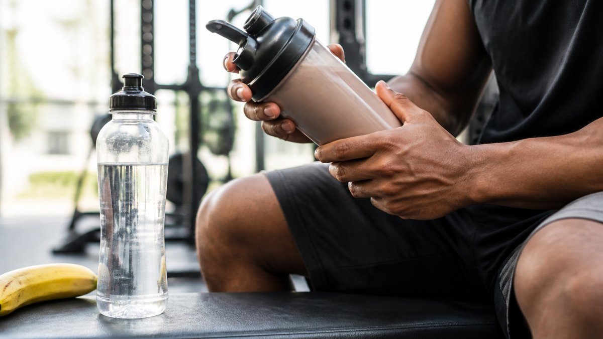 Close-up of hands shaking a plant-based protein powder in a shaker bottle on a gym bench, with a banana and water bottle nearby