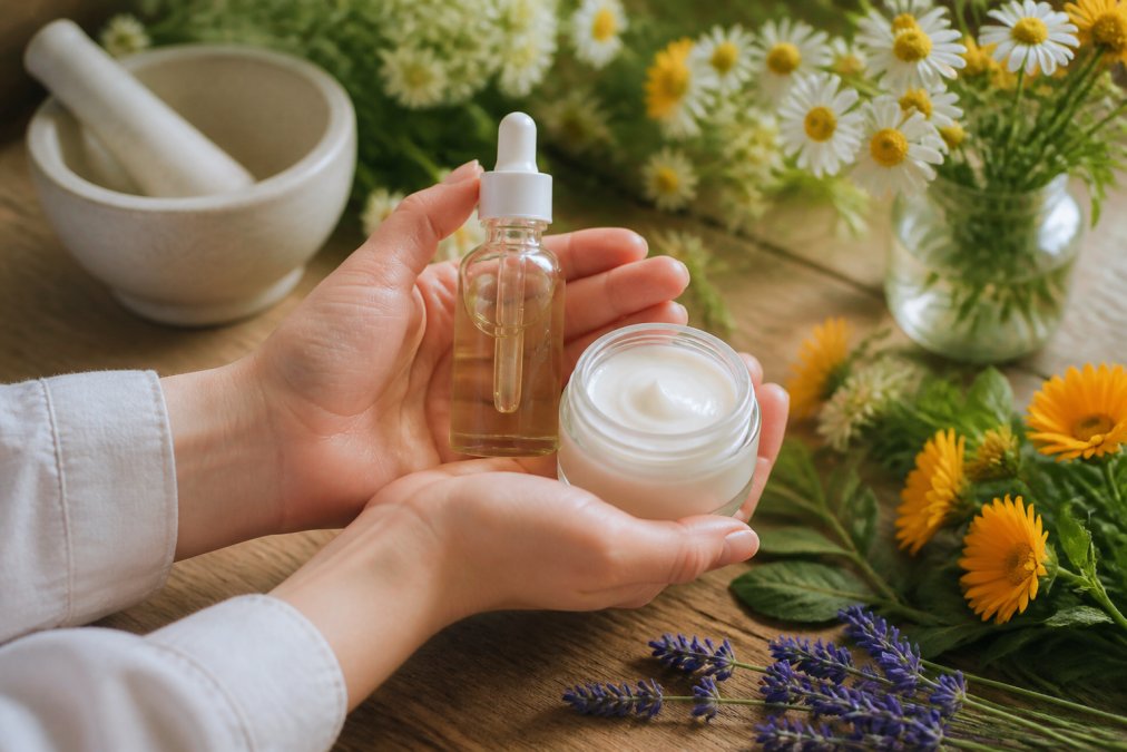 Nahaufnahme von Händen, die natürliche Hautpflegeprodukte in minimalistischen Glasflaschen halten, mit verschwommenen frischen Botanicals wie Kamillenblüten und Ringelblumen im Hintergrund