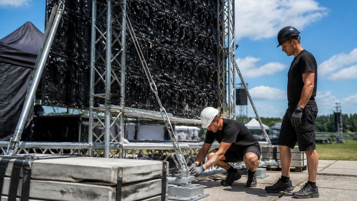 Een grote outdoor LED-wall gemonteerd op een truss-constructie tijdens een buitenevenement op een zonnige dag, met technici die de verankeringspunten en ballastgewichten controleren aan de voet van de constructie