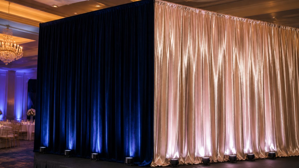 Professional event setup showing velvet drape on left side in deep jewel tone absorbing stage lighting and creating rich color depth, satin drape on right side reflecting ambient uplighting with visible glassy sheen, both in formal ballroom setting with theatrical lighting rigs