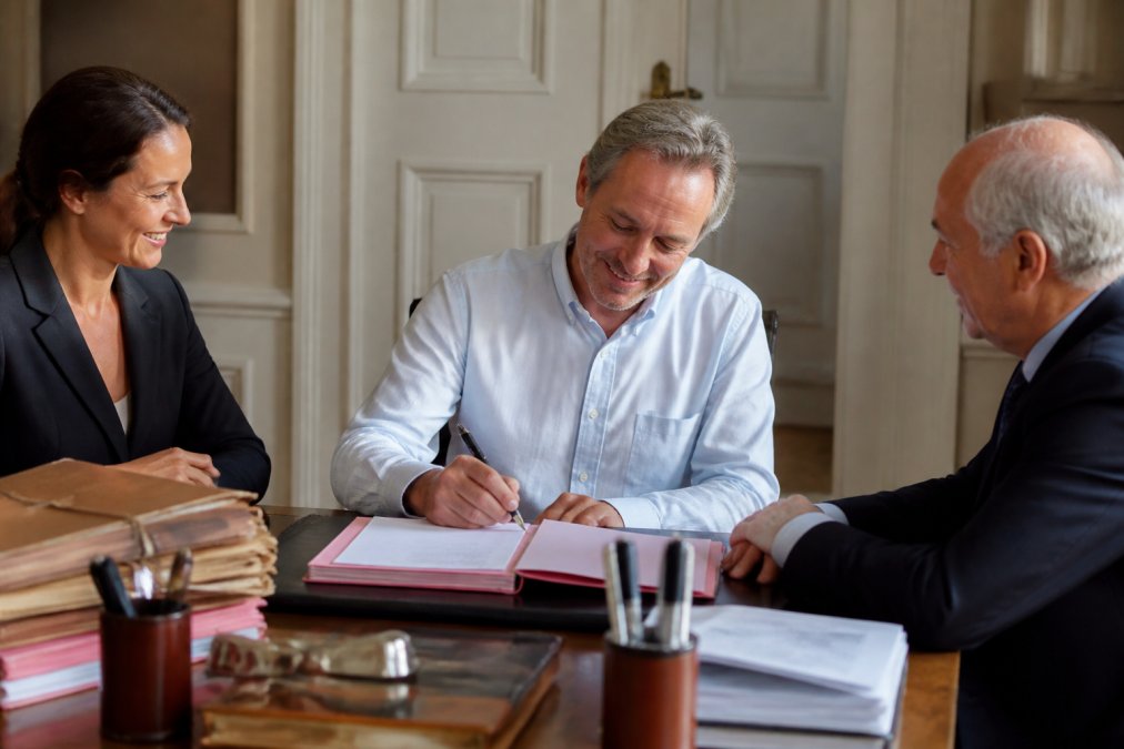 Propriétaire soulagé et souriant signant les documents finaux chez le notaire, entouré de dossiers organisés et de stylos, avec une expression de soulagement et de satisfaction