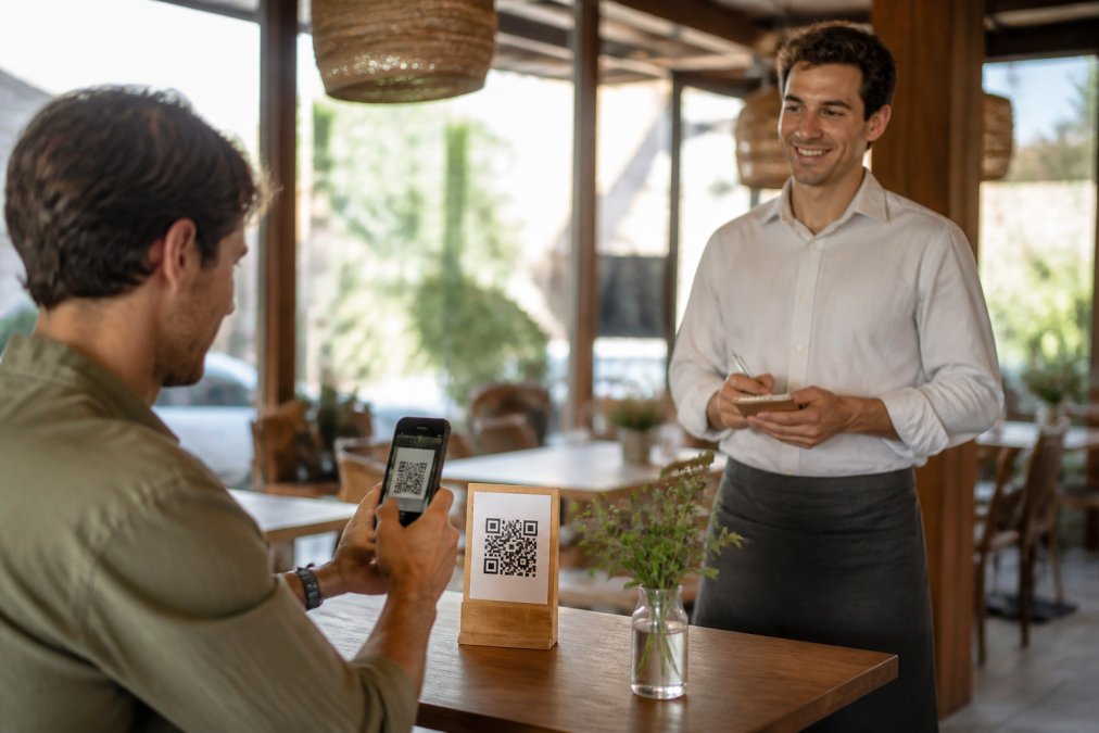 Un comensal escanea con su teléfono móvil un código QR colocado en un soporte de madera sobre la mesa de un restaurante luminoso, mientras el camarero espera sonriente con una libreta en la mano