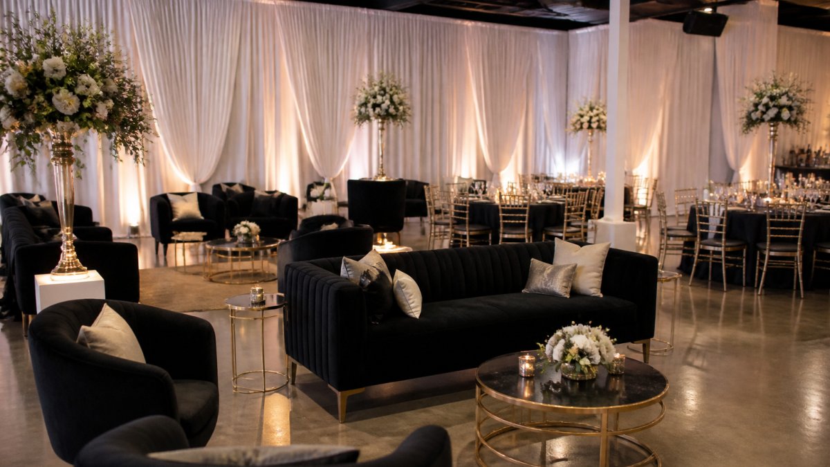 Elegant luxury wedding venue interior with sophisticated draping, professional lounge furniture arranged for cocktail reception, ambient up-lighting creating warm ambiance, and refined table settings