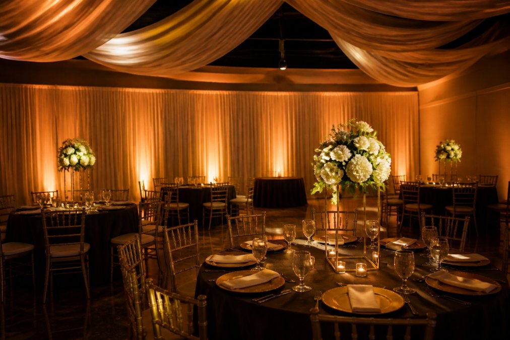 Elegant event venue interior with warm amber uplighting washing draped walls, pin spots illuminating floral centerpieces on round tables, and soft fabric ceiling draping overhead