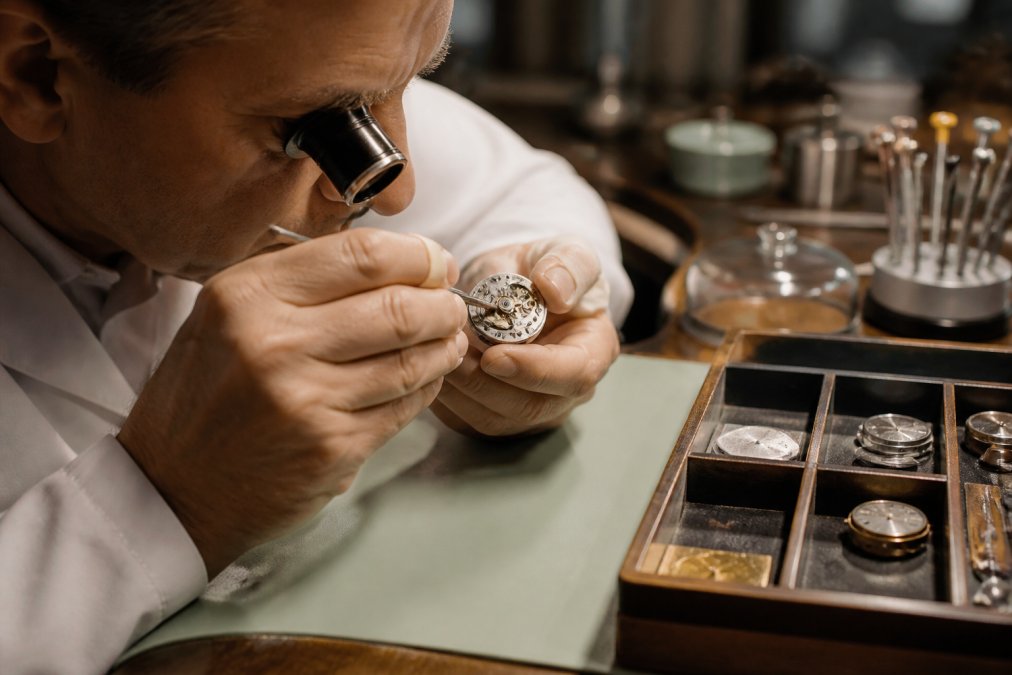 Watchmaker examining a mechanical movement under a jeweler's loupe in a workshop setting, with precision tools and component trays visible under warm task lighting