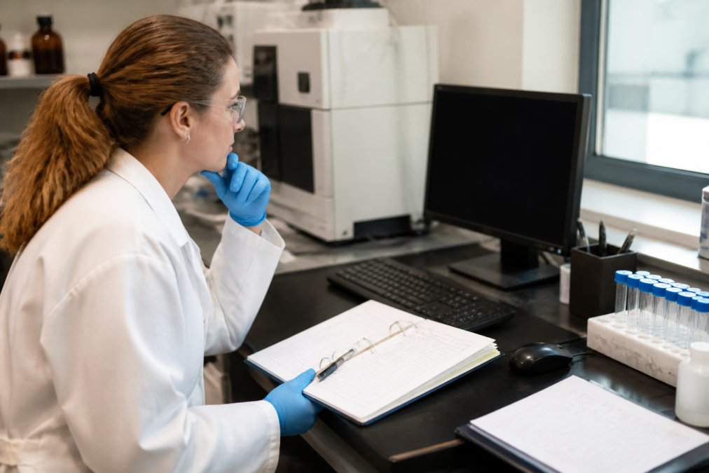 A scientist in a lab coat reviewing accreditation documents on a computer screen in a bright laboratory setting, with a clipboard and notes visible on the desk