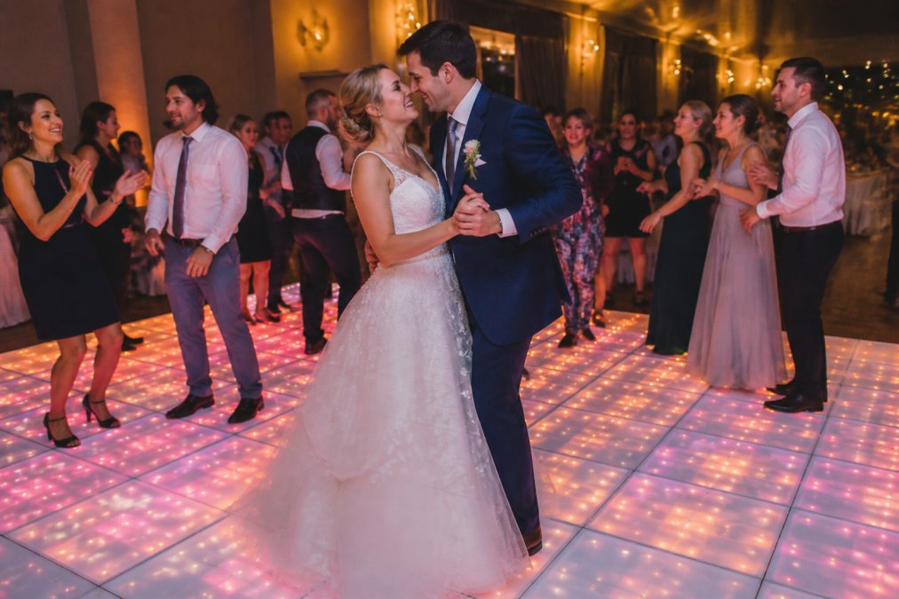 Bruidspaar danst op een verlichte led-dansvloer met zachte roze en gouden kleuren tijdens een avondbruiloftsfeest, gasten dansen op de achtergrond in feestkleding, warme sfeerverlichting in de zaal