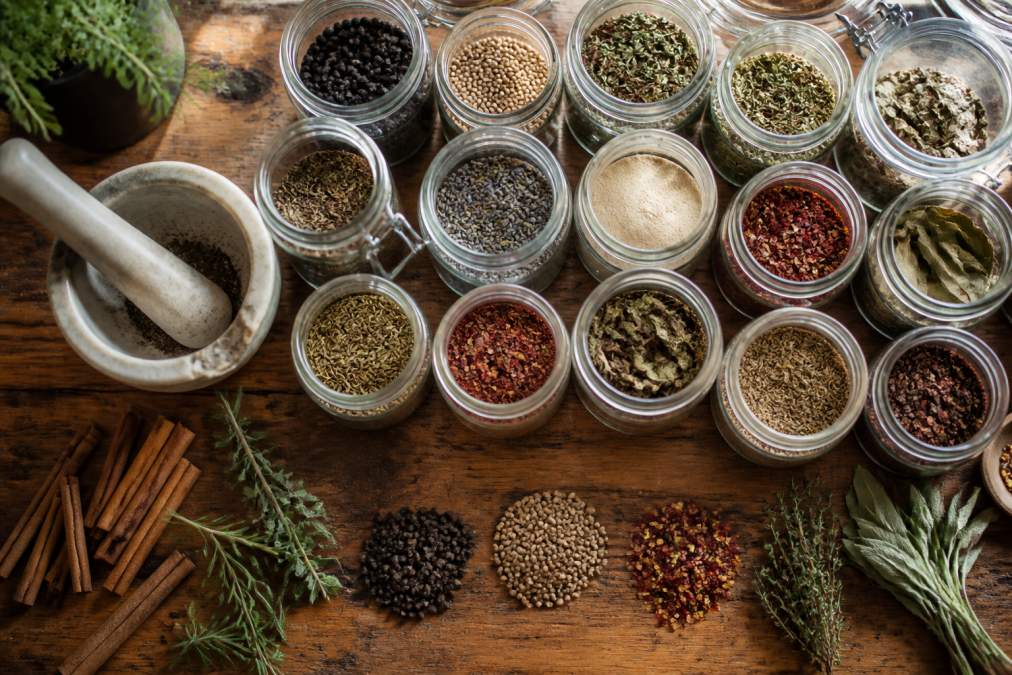 Overhead view of an organized spice collection in glass jars with handwritten labels, arranged on a wooden countertop next to a ceramic mortar and pestle, with dried herbs and whole spices visible, natural afternoon light illuminating the scene