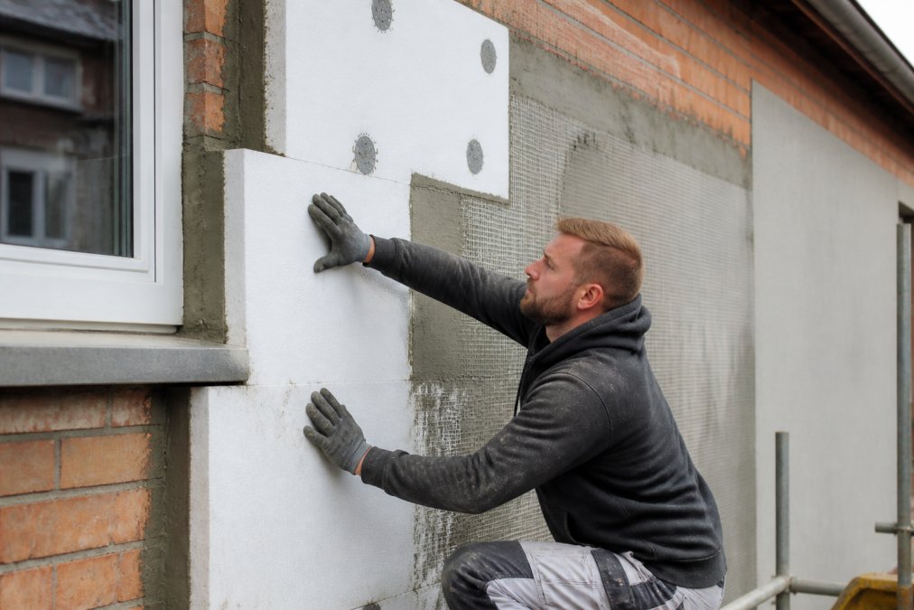Close-up van een vakman die isolatieplaten aanbrengt op de buitenmuur van een rijwoning, met zichtbare wapeningslaag en gevelafwerking in uitvoering