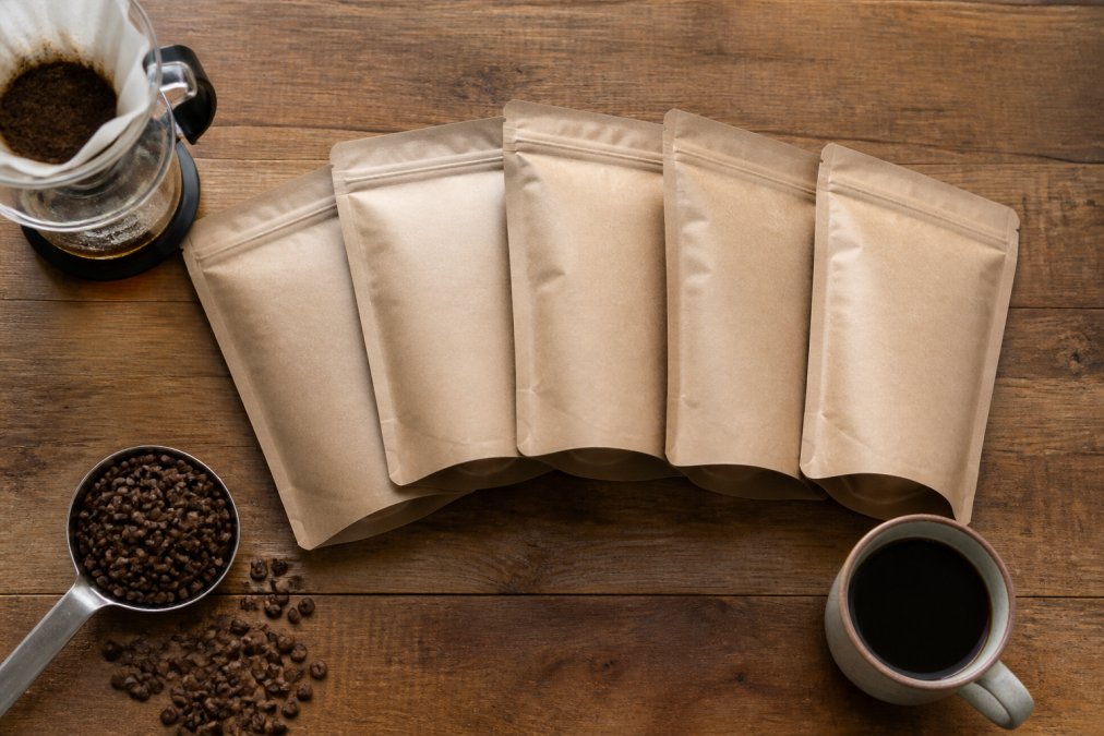 Flat lay of coffee bags, a pour-over dripper, whole beans, and a filled mug on a wooden kitchen counter.
