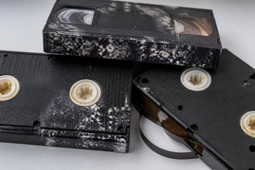 Close-up of deteriorating VHS tapes showing visible mould, discoloured spools, and tape warping on a white surface