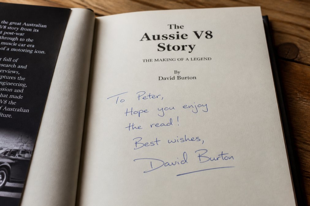 Close-up photograph of a book's title page showing an author's handwritten signature and personal inscription in blue ink, with the book's cover visible at the edge