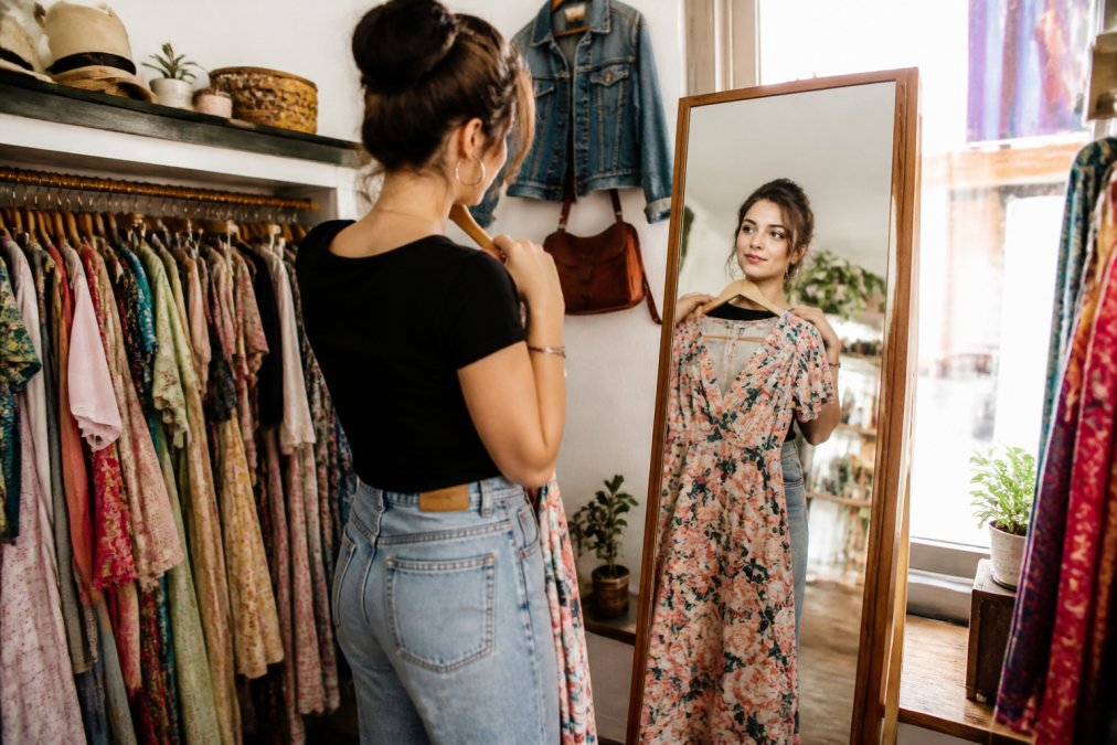 Woman browsing colorful vintage clothing in a sunlit boutique while holding a floral dress up to a mirror
