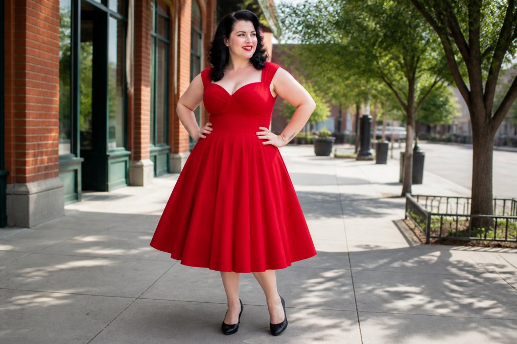 Woman with curvy figure wearing a red vintage-inspired swing dress with fitted waist and flared skirt, standing confidently with hands on hips, showing how the dress hugs curves at the waist and hips