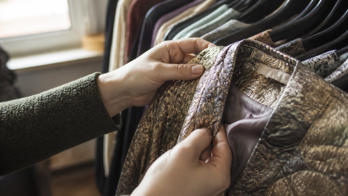 Nahaufnahme von Händen, die ein Vintage-Kleidungsstück detailliert untersuchen, Etiketten werden gelesen, Nähte genau inspiziert, Stoffstruktur unter natürlichem Licht sichtbar